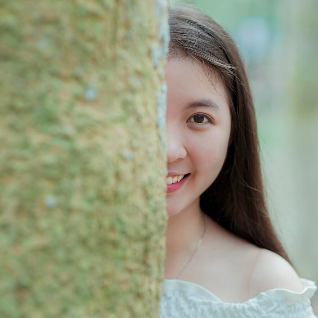 Happy young woman peeking from behind a tree, outdoors in nature.